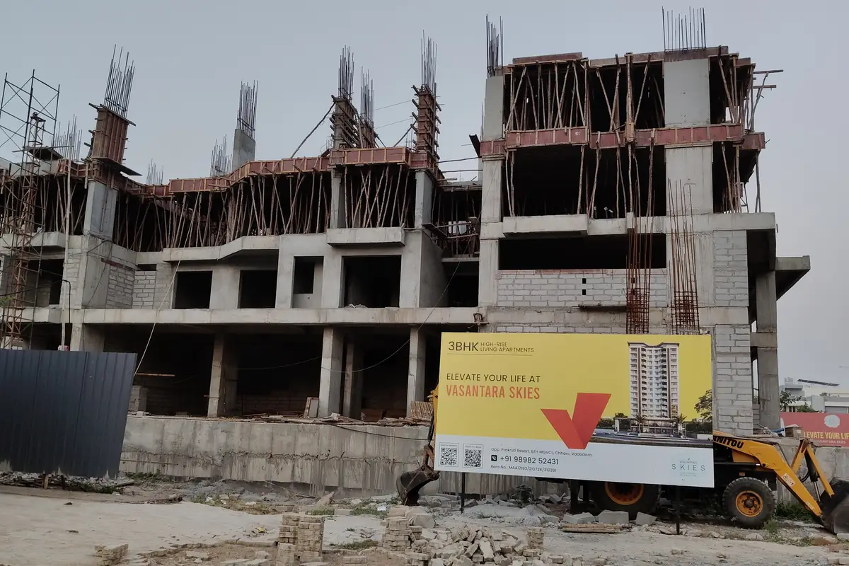 Vasantara Skies under construction — three slabs cast, columns rising, site hoarding on the road in Chhani, Vadodara